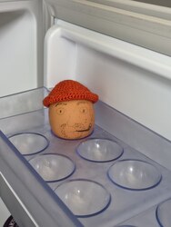 Un œuf coiffé d'un petit bonnet orange au crochet, posé dans le bac à œufs du frigo, avec un visage dessiné dessus